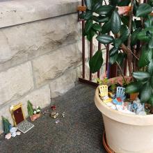 A small wooden door decorated with miniature trees, shells, and a birdbath. To the right, the soil in a planted pot is decorated with minature beach chairs and a sandcastle.