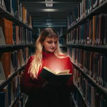 Woman standing in library stacks with a lit-up book.