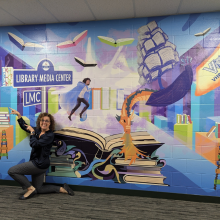 A woman kneels and gestures towards a wall mural filled with book and library imagery, books, a ship, and a dragon. 