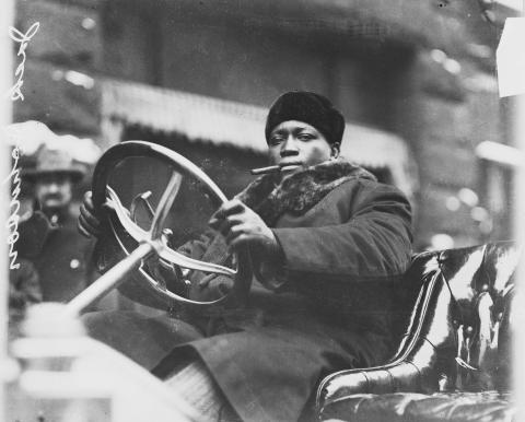 Black man sitting behind a wheel of an old convertible car with a cigar in his mouth