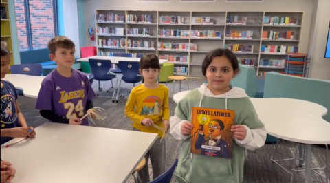 Four students around a table, showing a Lewis Latimer picture book, working on an invention in STEAM club.