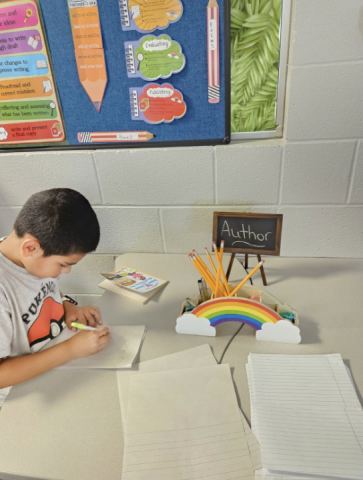 A young boy is sitting at a desk, writing on paper with a pencil.