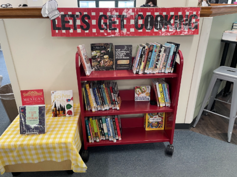 Library display with a red cart filled with various cookbooks, organized on three shelves. A banner above reads 'LET'S GET COOKING' in a red and white checkered pattern.