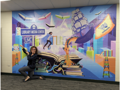 A woman kneels and gestures towards a wall mural filled with book and library imagery, books, a ship, and a dragon. 